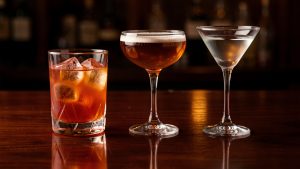 Trois cocktails classiques alignés sur un comptoir en bois sombre, avec un Negroni orangé sur glace, un Manhattan ambré en coupe et un Dry Martini transparent, baignés dans une lumière de bar élégante.