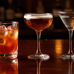 Trois cocktails classiques alignés sur un comptoir en bois sombre, avec un Negroni orangé sur glace, un Manhattan ambré en coupe et un Dry Martini transparent, baignés dans une lumière de bar élégante.